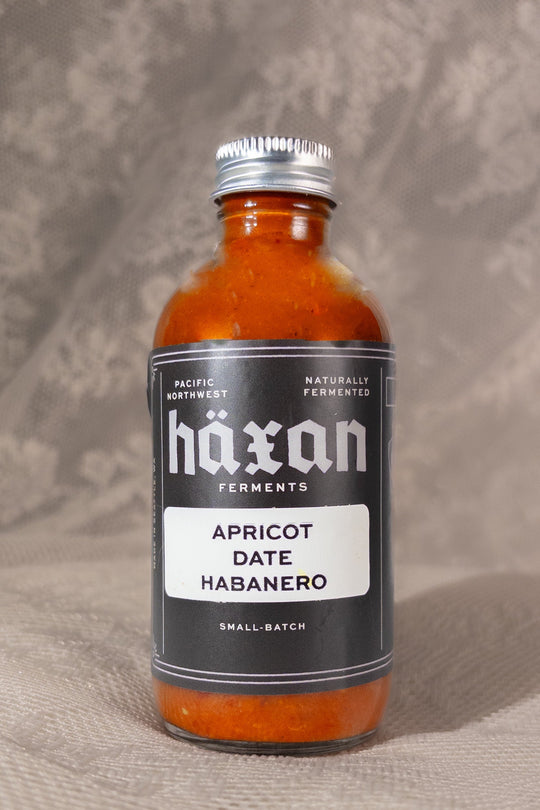 A small glass bottle of Haxan Ferments Apricot Date Habanero sauce, showing the vibrant orange-red sauce inside, with a black label and silver cap, set against a textured neutral background.
